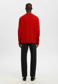Mann steht mit dem Rücken zugewandt, trägt einen roten Pullover, schwarze Jeans und schwarze Schuhe vor einem schlichten hellen Hintergrund.