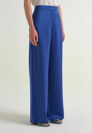 Donna con pantaloni a vita alta, a gamba larga blu reale e tacchi aperti blu, in piedi contro uno sfondo chiaro e semplice.