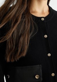 Langt brunt hår over en sort ribbet cardigan med sølvfarvede trykknapper og en lomme på brystet.