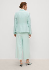 Mintgroene pakken met een maatjasje en wijde broekspijpen, gemaakt van gladde stof. Heeft een getailleerde taille en een enkele achternaad detail.