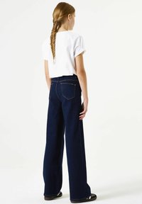 Blauwe denim broek met wijde pijpen en een hoge taille, voorzien van oranje stiksel details en klassieke achterzakken. Gestyled met een wit topje met korte mouwen.