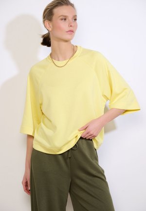 Young woman wearing a loose yellow short-sleeve shirt, olive green pants, and a gold chain necklace, posing against a white background.