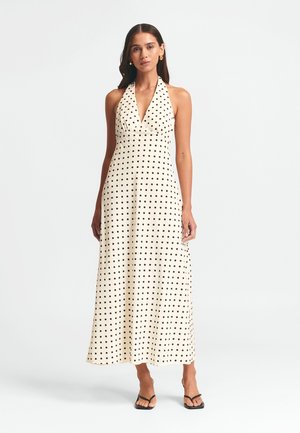 Halenková maxi šaty s puntíky v krémové barvě s černými tečkami, s V-výstřihem a splývavou látkou. Na sobě má černé sandály.