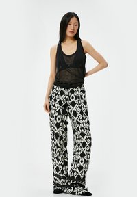 Zwarte doorschijnende tanktop met knoopdetails, gecombineerd met hoogriem wijdvallende broek met zwart-wit abstract patroon en franjes.