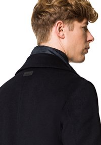 Czarny wełniany płaszcz o teksturowanym wykończeniu, z wyraźnym kołnierzem i gładką podszewką. Szczegół z logo na tylnym obszarze szyi.