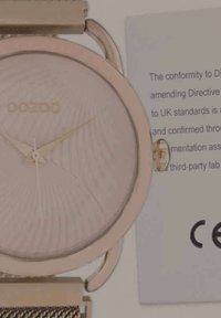 Montre-bracelet en or rose avec un cadran texturé, des aiguilles fines et un bracelet en maille métallique ; inclut une étiquette de conformité CE à côté.