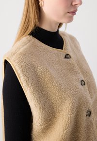 Beige, fluffig lammullsväst med rundad halsringning och svarta knappar. Bärs över en svart, figursydd polo. Har råa kanter och en mjuk textur.