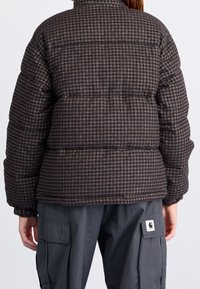 Doudoune à carreaux marron avec un col haut et des poignets élastiques. Motif matelassé épais et patch logo sur le côté gauche d'un pantalon gris.