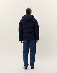 Mann iført marineblå dunjakke med hette, blå jeans, sorte sko og en svart baseballcaps, står med ryggen til foran en ensfarget bakgrunn.