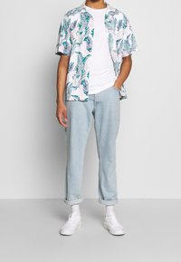 Kortärmad skjorta med knappar i vitt med grön och blå bladtryck. Bärs över en enkel vit t-shirt, ihop med ljusblå jeans och vita sneakers.