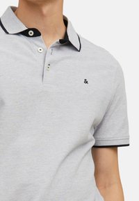 Lichtgrijze polo met een zwarte kraag, een knoopsluiting met drie knopen en een klein geborduurd logo op de borst. Zachte, gestructureerde stof.