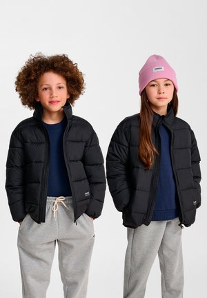 Zwei Kinder in schwarzen Daunenjacken, marineblauen Hemden und grauen Jogginghosen stehen nebeneinander, eines trägt eine pinke Mütze, beide haben die Hände in den Taschen.