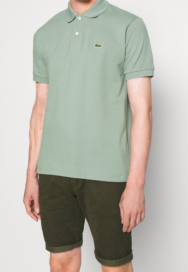Homme portant un polo vert sauge à manches courtes avec un petit logo crocodile et un short longueur genou couleur olive foncée retroussé.