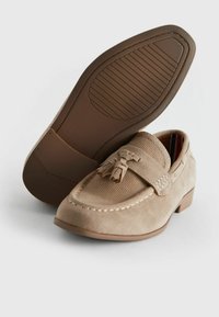 Mocassins en suédine beige avec une tige texturée, des glands décoratifs et une semelle en caoutchouc marron basse avec un dessous à motif antidérapant.