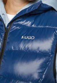 Person som bär en blå vadderad väst med "HUGO"-logotyp över en ljusblå hoodie, delvis synlig dragkedja och nacke.