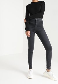 Osoba v černých skinny džínách s vysokým pasem, černém dlouhém rukávu a bílých teniskách stojící proti bílému pozadí.