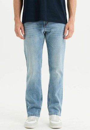 Mann trägt hellblaue Jeans mit geradem Schnitt, weiße Sneaker und ein dunkelblaues Hemd, steht vor einem einfarbigen Hintergrund.