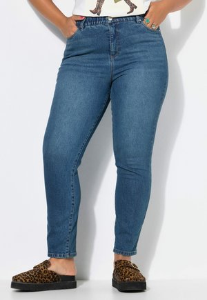Frau trägt blaue Slim-Fit-Jeans mit Händen in den Taschen und Leopardenmuster-Slip-ons, steht vor weißem Hintergrund.