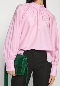 Blusa rosa chiaro con colletto alto e dettagli arricciati, abbinata a una borsa a spalla verde con texture coccodrillo e pantaloni neri.