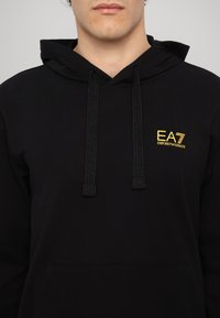 Black hoodie made of cotton blend featuring a drawstring hood, kangaroo pocket, and a gold logo with "EA7 EMPORIO ARMANI" on the chest.