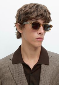 Sunglasses med en genomskinlig brun ram och mörka linser, med sköldpaddsaccenter. Bärs med en brun skjorta med krage och ett rutigt blazer.