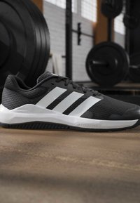 Chaussure de sport noir et blanc avec trois bandes blanches sur le côté, posée sur un sol de salle de sport avec du matériel de musculation en arrière-plan.