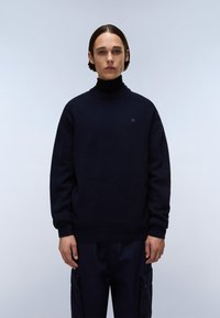 Navyblauer Strickpullover mit einem gerippten Rollkragen, versehen mit einem kleinen Logo auf der Brust und lässigen Ärmeln, getragen mit dunklen Cargo-Hosen.