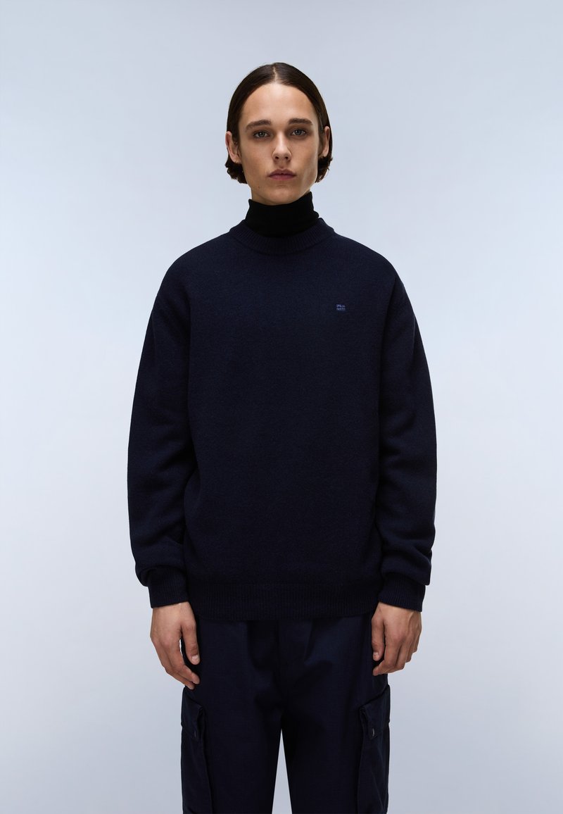 Navyblauer Strickpullover mit einem gerippten Rollkragen, versehen mit einem kleinen Logo auf der Brust und lässigen Ärmeln, getragen mit dunklen Cargo-Hosen.