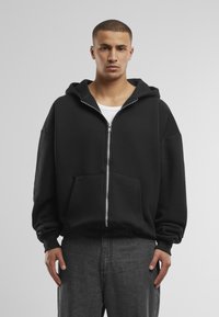 Schwarze Zip-Hoodie mit großer Fronttasche, Kapuze mit Kordelzug und lässiger Passform. Getragen über einem weißen Shirt, kombiniert mit dunkelgrauen Jeans.