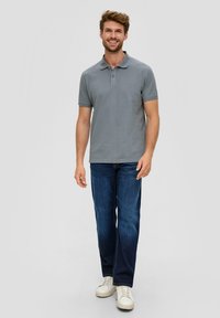 Polo shirt gris à manches courtes avec un col, associé à un jean bleu foncé et des baskets blanches, debout sur un fond uni.