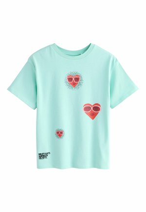 Lysegrøn mintfarvet bomuldst-shirt med røde hjertegrafikker iført solbriller. Sort tekst nederst siger "DOLLFANTS BETWEEN HEARTS."