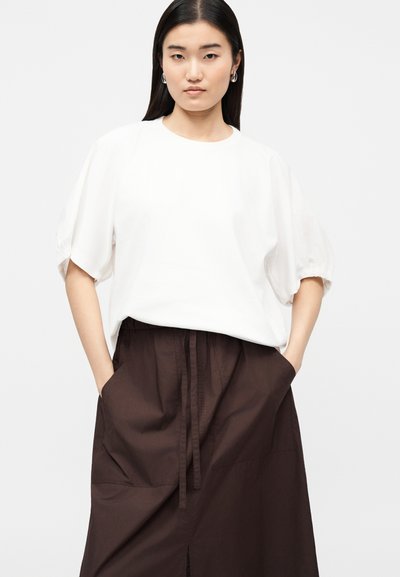 Γυναίκα με μακριά σκούρα μαλλιά που φορά ασημένια σκουλαρίκια κρίκους, μια φαρδιά λευκή μπλούζα με κοντά μανίκια και φούστα ψηλόμεση σκούρου καφέ χρώματος με τσέπες.