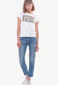 Balts T-krekls ar pelēku grafisku apdruku, saskaņots ar gaiši zilu džinsa biksēm. Modelis valkā baltas sporta kurpes. Vienkārša silueta, va�ļīga piegriezuma.