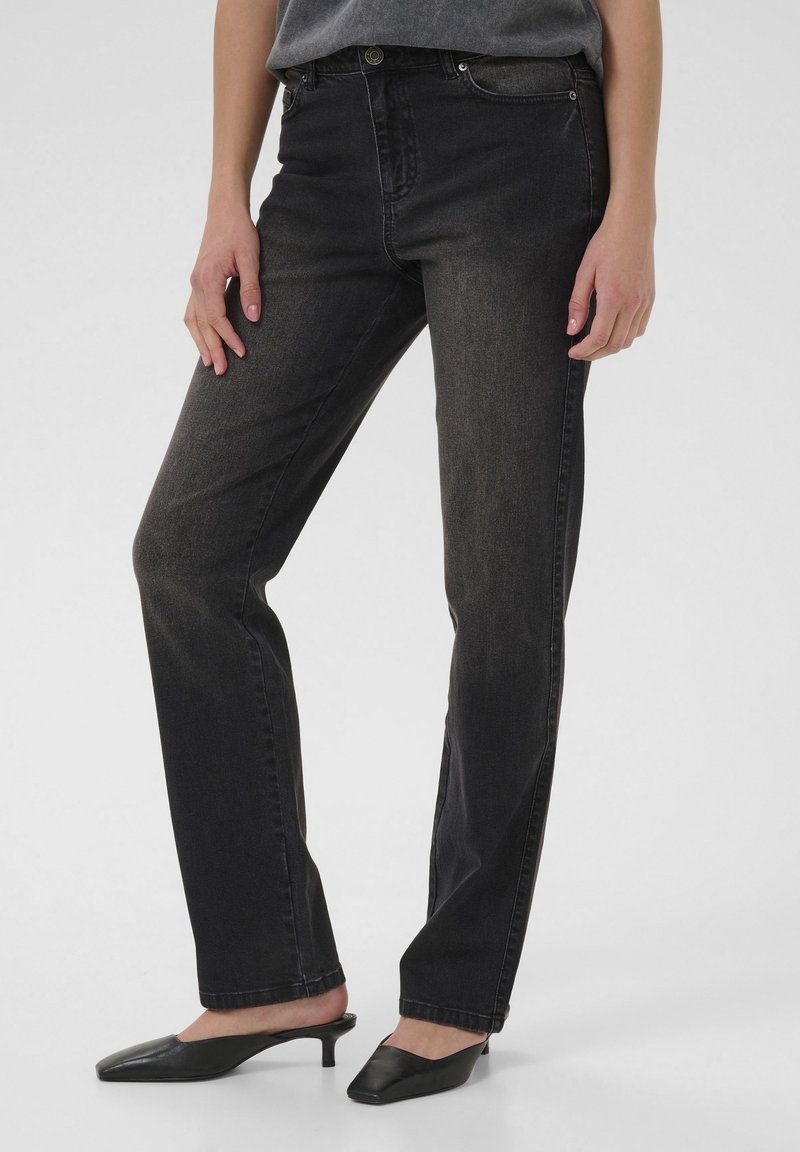 Personne portant un jean noir à jambes droites et des mules noires à talon bas pointu, se tenant devant un fond clair uni.