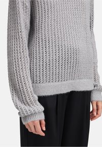 Hellgrauer, gestrickter Pullover mit offenem Maschenmuster, lässiger Passform, gerippten Bündchen und weicher Textur. Kombiniert mit schwarzen Hosen.