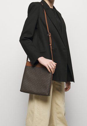 Personne portant un blazer noir et un pantalon beige, tenant un sac bandoulière marron à motifs avec une sangle en cuir sur fond blanc.