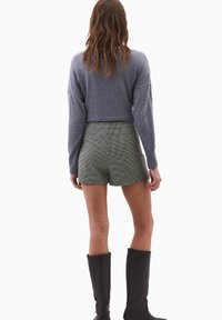 Kvindelig person står med ryggen til kameraet iført en grå cropped sweater, ternede høj talje shorts og sorte knæhøje støvler.
