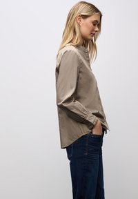 Camicia beige con colletto alla mandarina, maniche lunghe e orlo arrotondato. Abbinata a jeans blu scuro, che mostrano un fit casual.