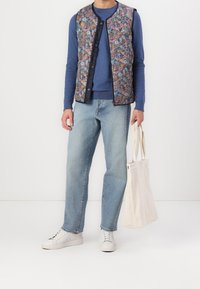 Väst med blommönster och vaddering i marinblå bas, överlagrad på en blå tröja, kombinerad med ljusblå jeans och vita sneakers. Inkluderar en canvasväska.