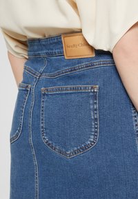 Gros plan d'une jupe en denim bleu avec poches arrière et étiquette en cuir beige SeeByChloé, portée avec un haut crème.