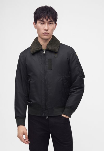 Jeune homme portant une veste bomber noire avec un col en peau de mouton vert foncé et un pantalon noir, debout devant un fond uni.