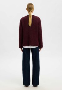 Burgunderfarbener Wollpullover mit lockerer Passform, langen Ärmeln und geripptem Saum, kombiniert mit weiten, dunklen Denim-Jeans. Rückansicht.