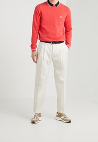 Roter Pullover mit Details am Kragen in Schwarz und Weiß, weiße plissierte Hosen und beigefarbene und weiße Sneaker. Glatte Texturen und lässige Passform.