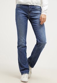 Sinisen denimin suorat housut, joissa on viisi taskua ja hienovarainen kuluminen. Yhdistettynä valkoisiin lenkkareihin ja vaaleaan neuleeseen.