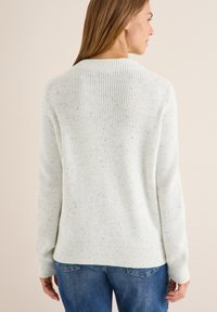 Donna con capelli lunghi di spalle, che indossa un maglione a maglia bianco con macchie e jeans blu, su uno sfondo neutro.
