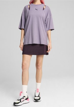 Fioletowa oversize'owa koszulka z wycięciami na ramionach i czarnym logo; w zestawie z czarną teksturowaną spódnicą i wielokolorowymi sneakersami.