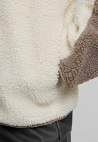 Sudadera de forro polar que presenta paneles en crema y marrón con una superficie texturizada y esponjosa. Puños elásticos y dobladillo en color neutro.