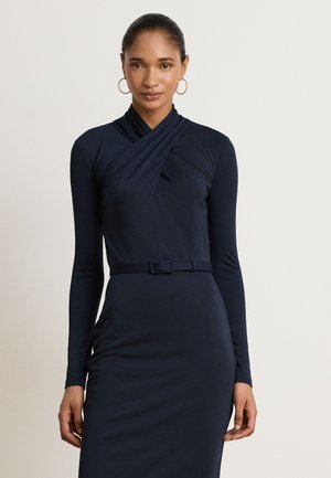 Femme aux cheveux tirés en arrière, portant des boucles d’oreilles créoles dorées et une robe bleu marine ajustée à manches longues, avec un décolleté croisé et une ceinture assortie.