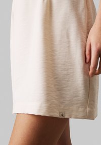 Abito in jersey di cotone rosa chiaro con una texture liscia, dotato di un'etichetta sul fondo con il logo "ck" sul lato inferiore, aderente in vita.