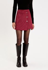 Bordowa zamszowa spódnica mini o kształcie A, z trzema srebrnymi guzikami z przodu, w zestawie z czarnymi kozakami do kolan.